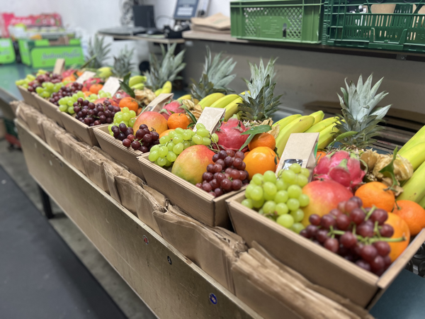 KI generiert: Kisten mit frischem Obst wie Trauben, Ananas und Äpfeln auf einem Tisch.
